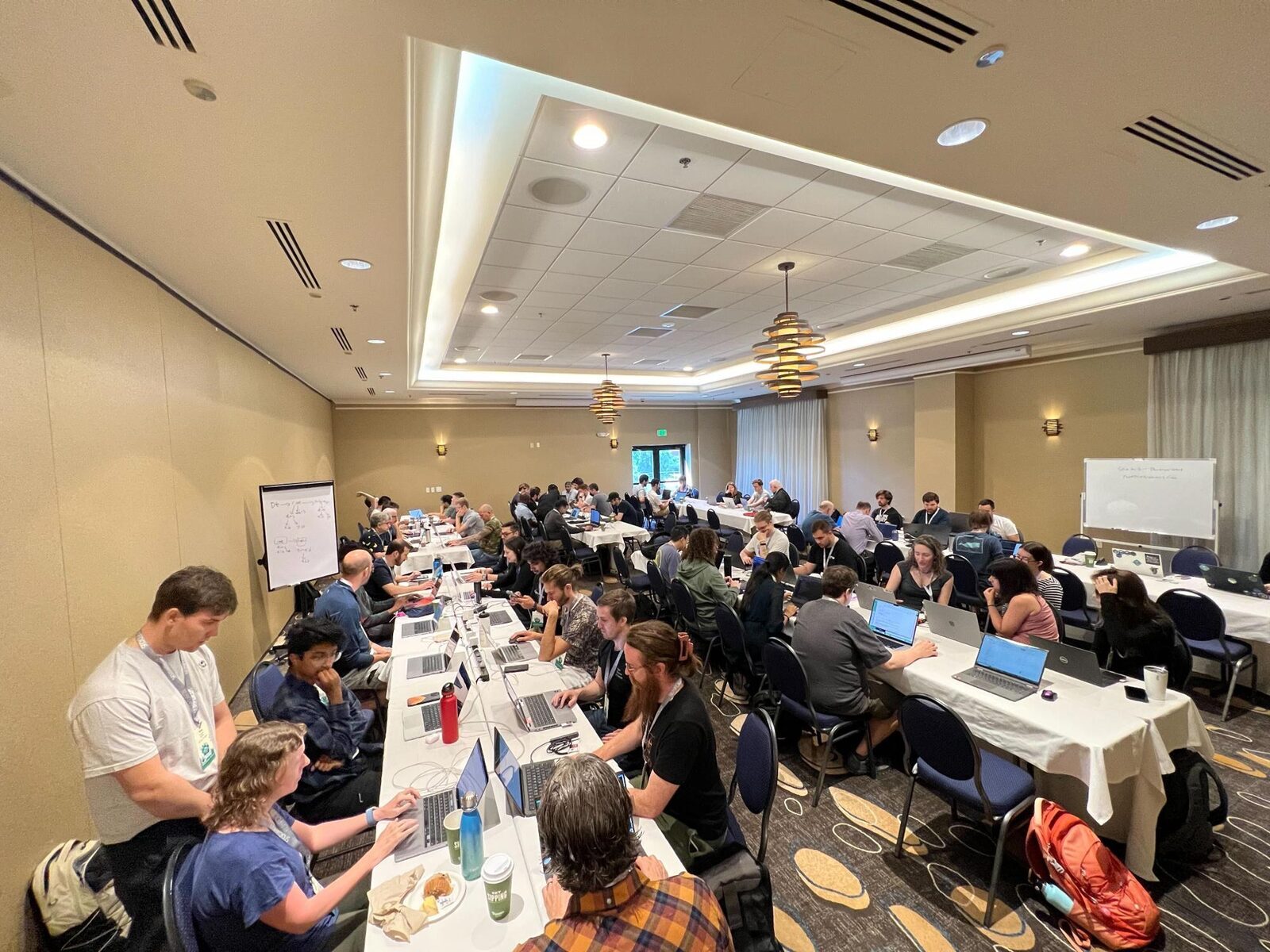 Photograph showing a tan room filled with long tables and people sitting in front of laptops working together on specific projects or tasks.